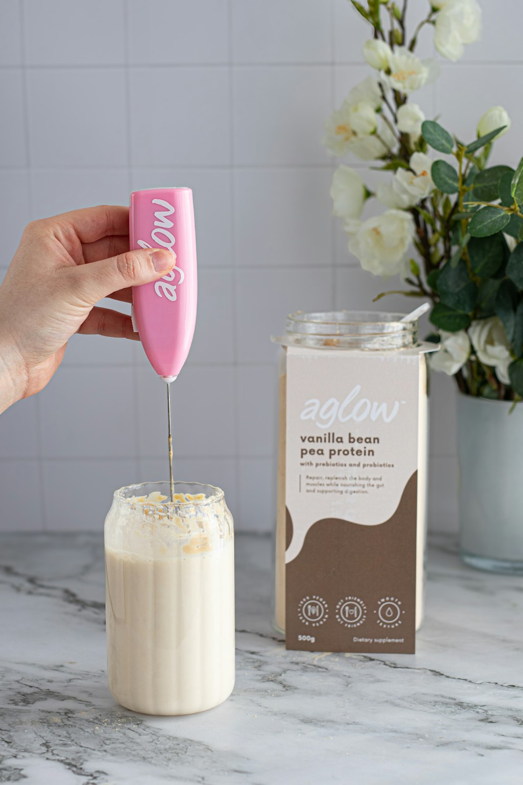 A hand holds a pink aglow handheld frother above a glass of creamy beverage, with the aglow vanilla bean pea protein powder jar standing beside it on a marble surface against a tiled wall and flowers in the background.